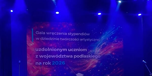 Scena oświetlona niebieskim światłem. Na ekranie znajduje się kolorowa grafika z napisem: ‘Gala wręczenia stypendiów w dziedzinie twórczości artystycznej uzdolnionym uczniom z województwa podlaskiego na rok 2026’