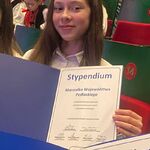 Dziewczynka siedząca na widowni trzyma otwartą teczkę z dyplomem Stypendium Marszałka Województwa Podlaskiego